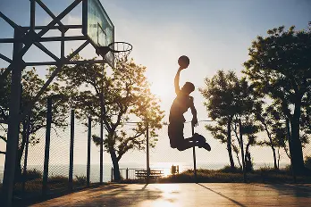 Jogador de basquetebol a realizar um afundanço num campo exterior ao pôr do sol, criando uma silhueta dramática contra o céu da noite.